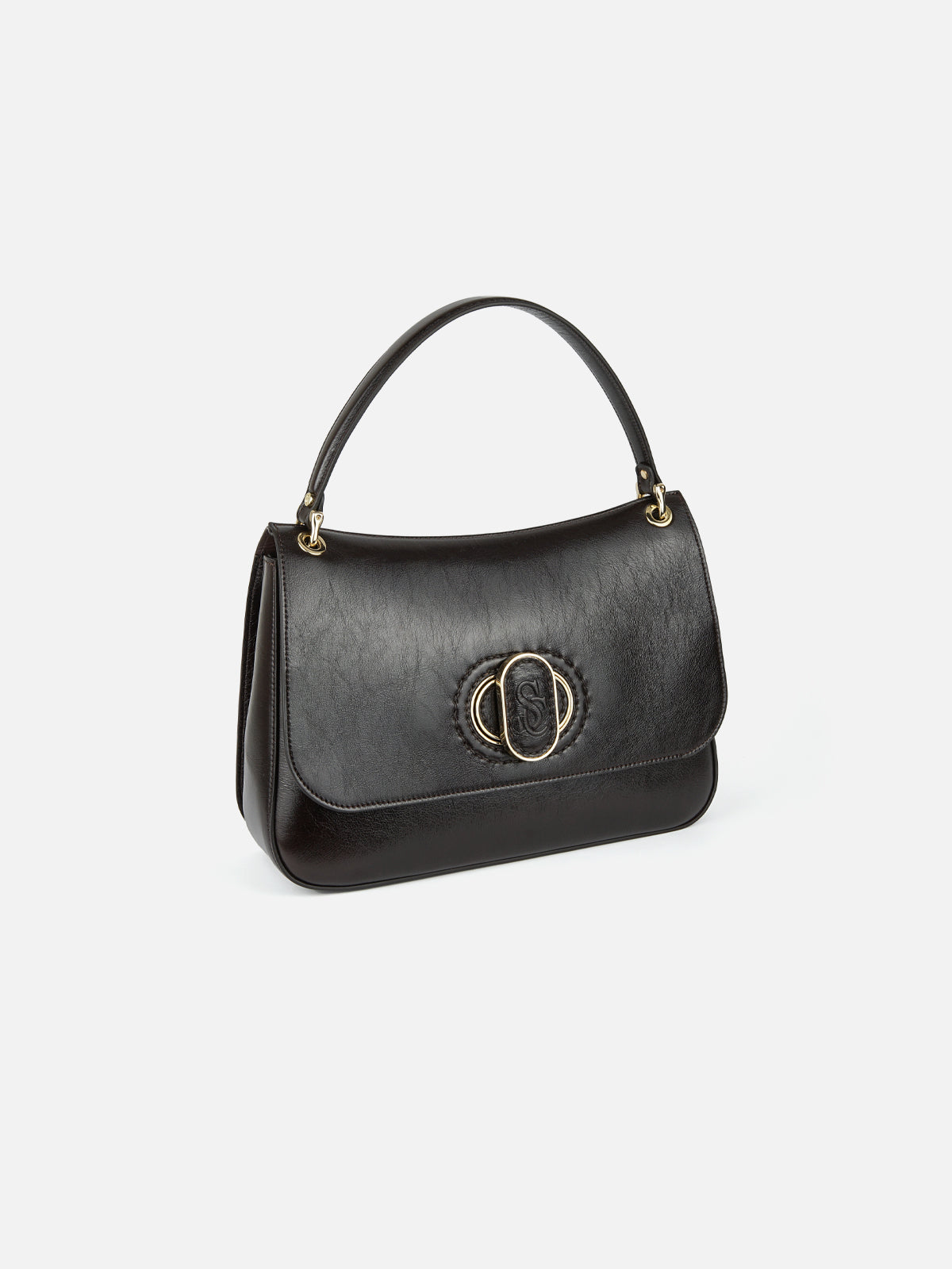 Three-quarter view of a black, slightly textured leather flap handbag. The bag has a single top handle attached with gold links and a central decorative gold oval hardware piece with an integrated logo. The background is a clean, plain light gradient.