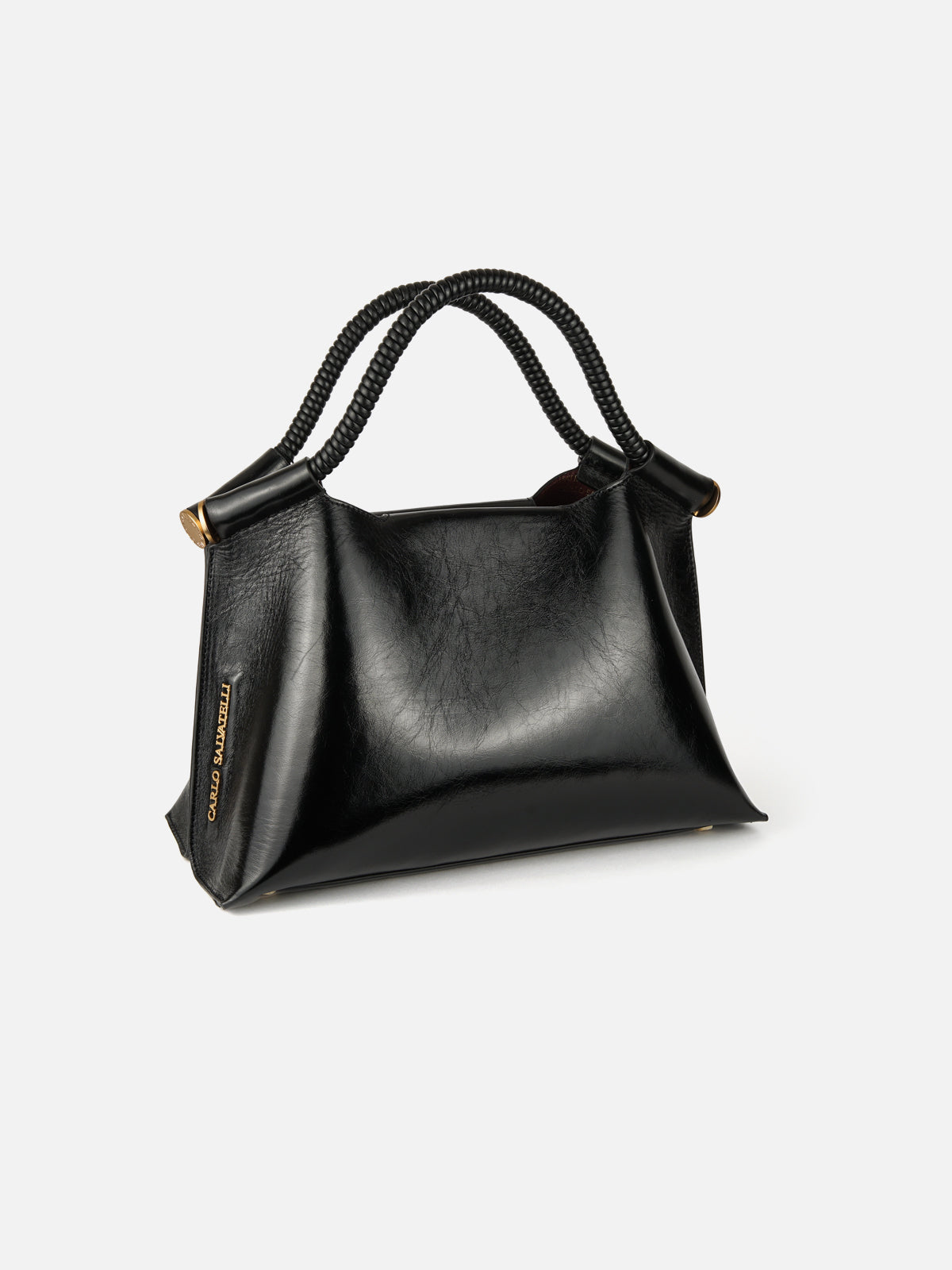 A three-quarter view of a structured, high-gloss black patent leather handbag featuring distinctive twisted black cord top handles. The bag has sculptural folds at the corners, polished gold-tone hardware connectors, and a vertical metal logo plate on the lower left side.